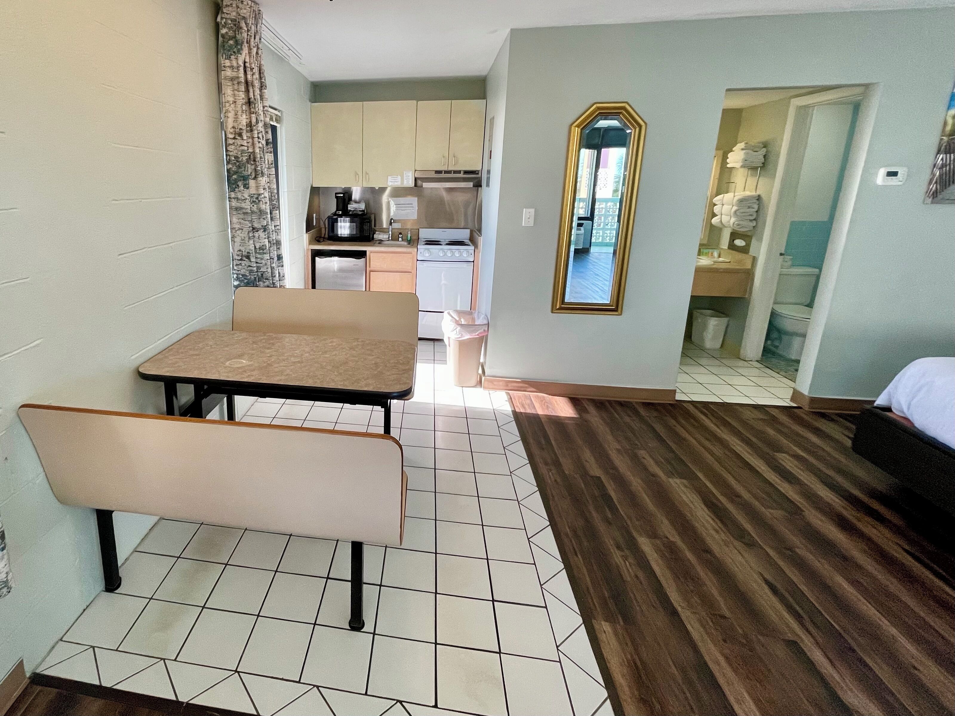 A compact hotel room with a kitchenette, a dining area with a table and bench, a mirror, and a bed.