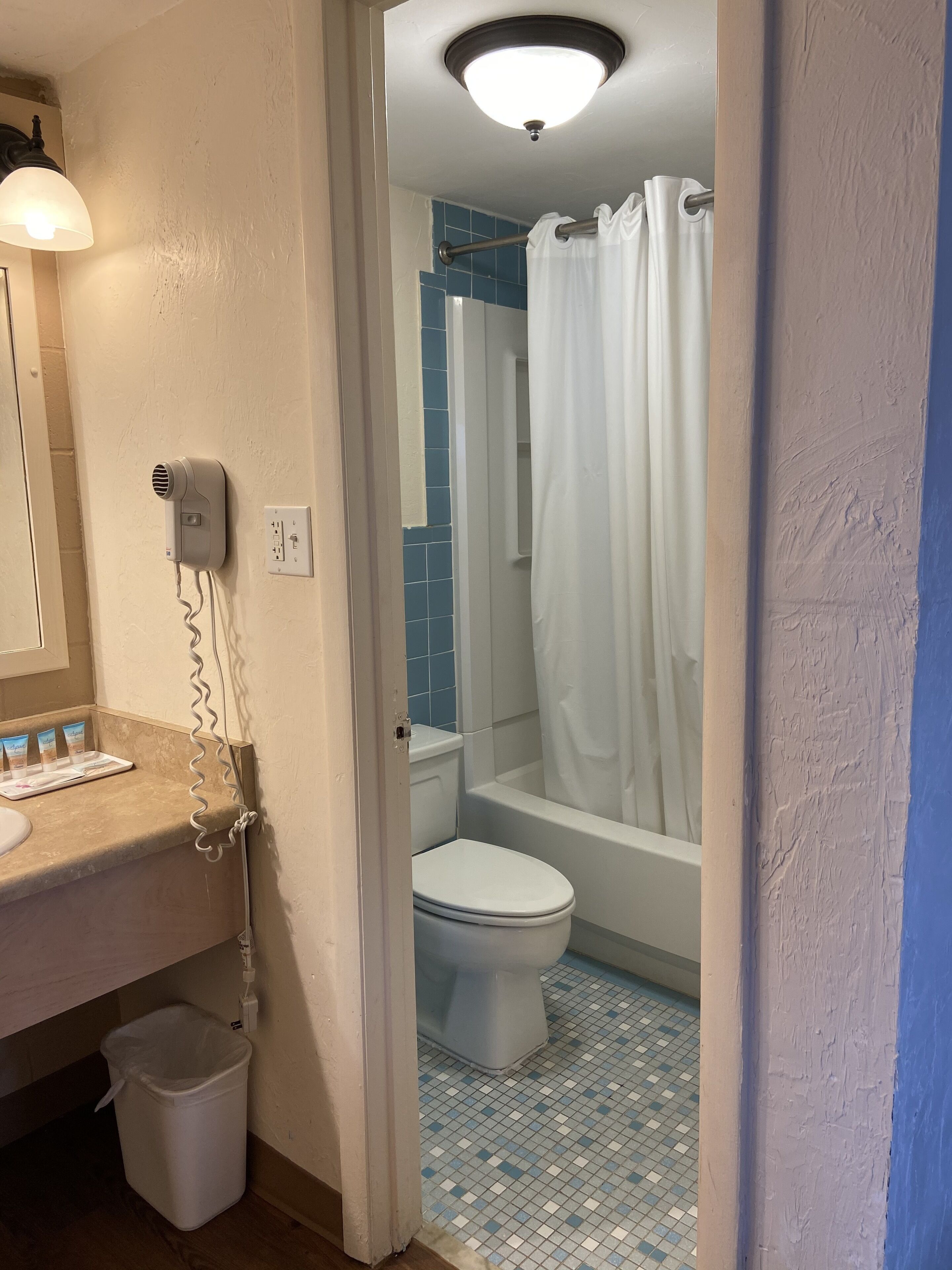 A bathroom with a toilet, a shower with a white curtain, a hairdryer on the wall, and a trash bin.