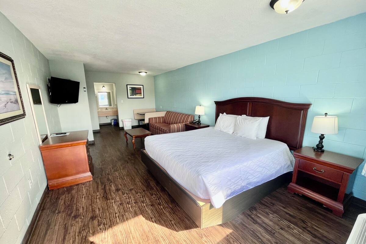 Una habitación de hotel con una cama grande, una mesita de noche de madera, un escritorio, una silla, un sofá, un televisor y un baño con espejo y lavamanos.