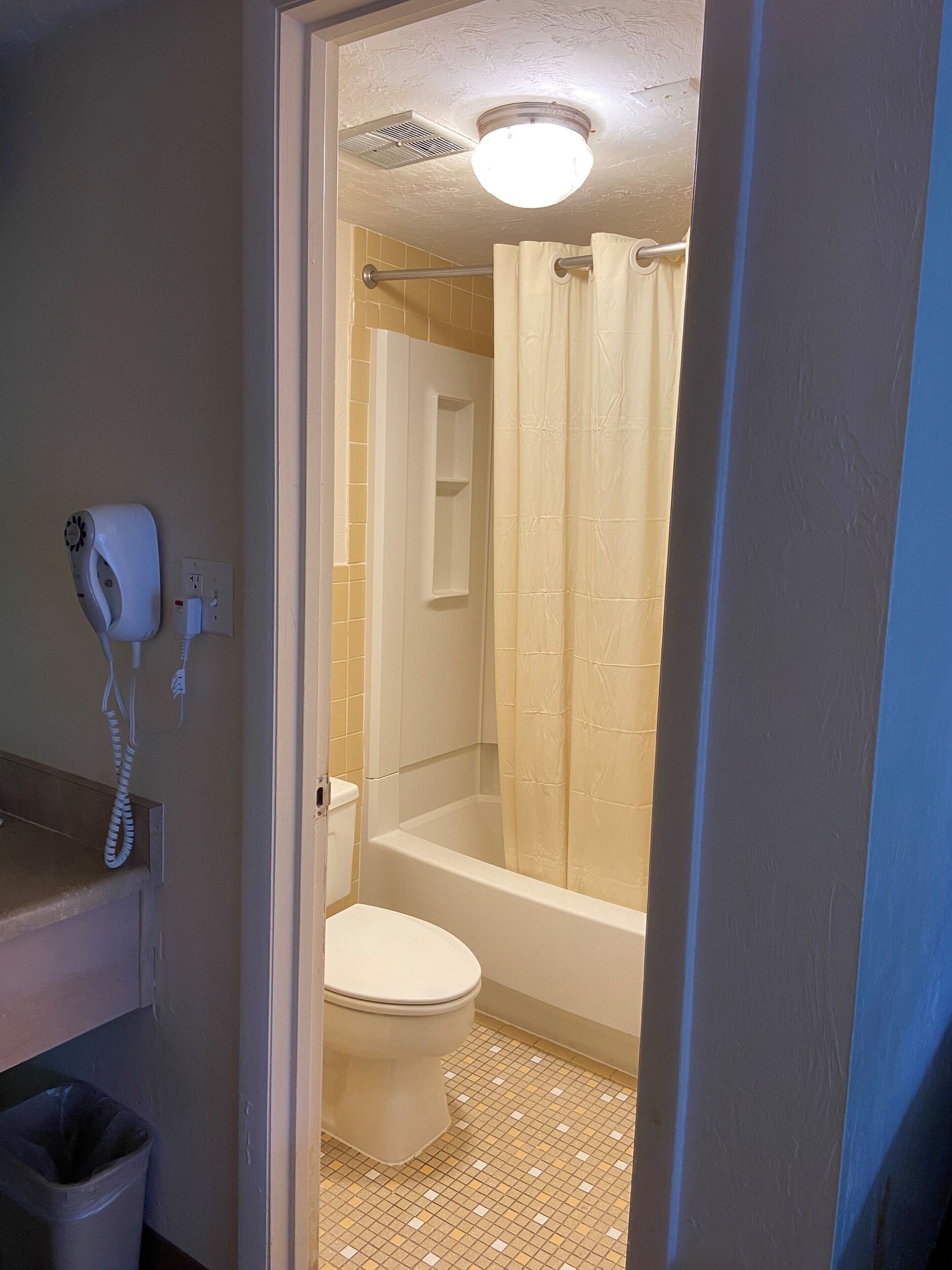 Une salle de bain comprenant des toilettes, une douche avec un rideau, un téléphone fixé au mur et un sol carrelé.