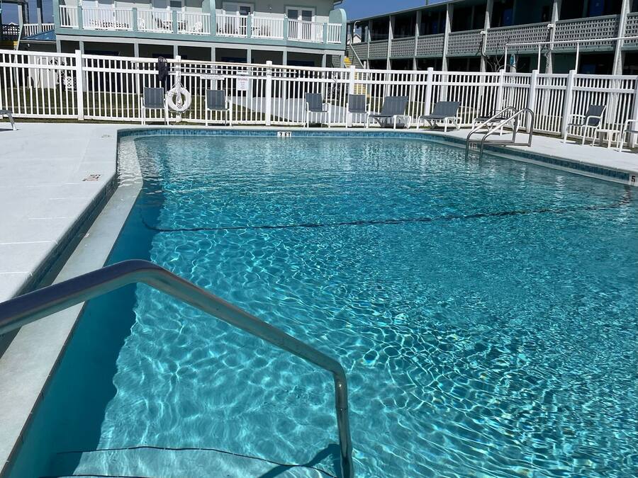 Una piscina cristalina con una escalera metálica, rodeada por una cerca blanca y un edificio con techo azul al fondo.