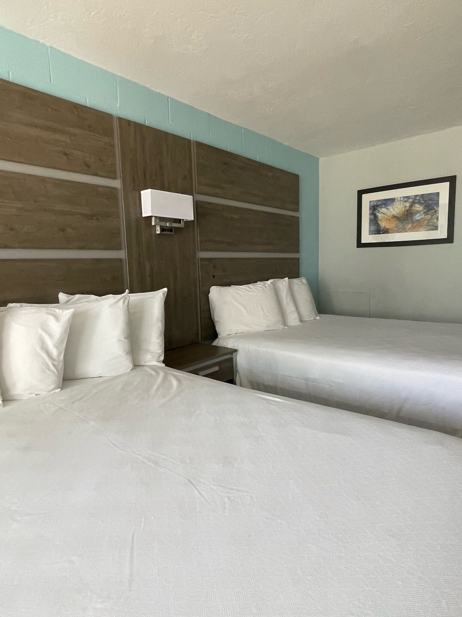 Habitación de hotel con dos camas, una mesita de noche, una lámpara de pared y un cuadro enmarco.