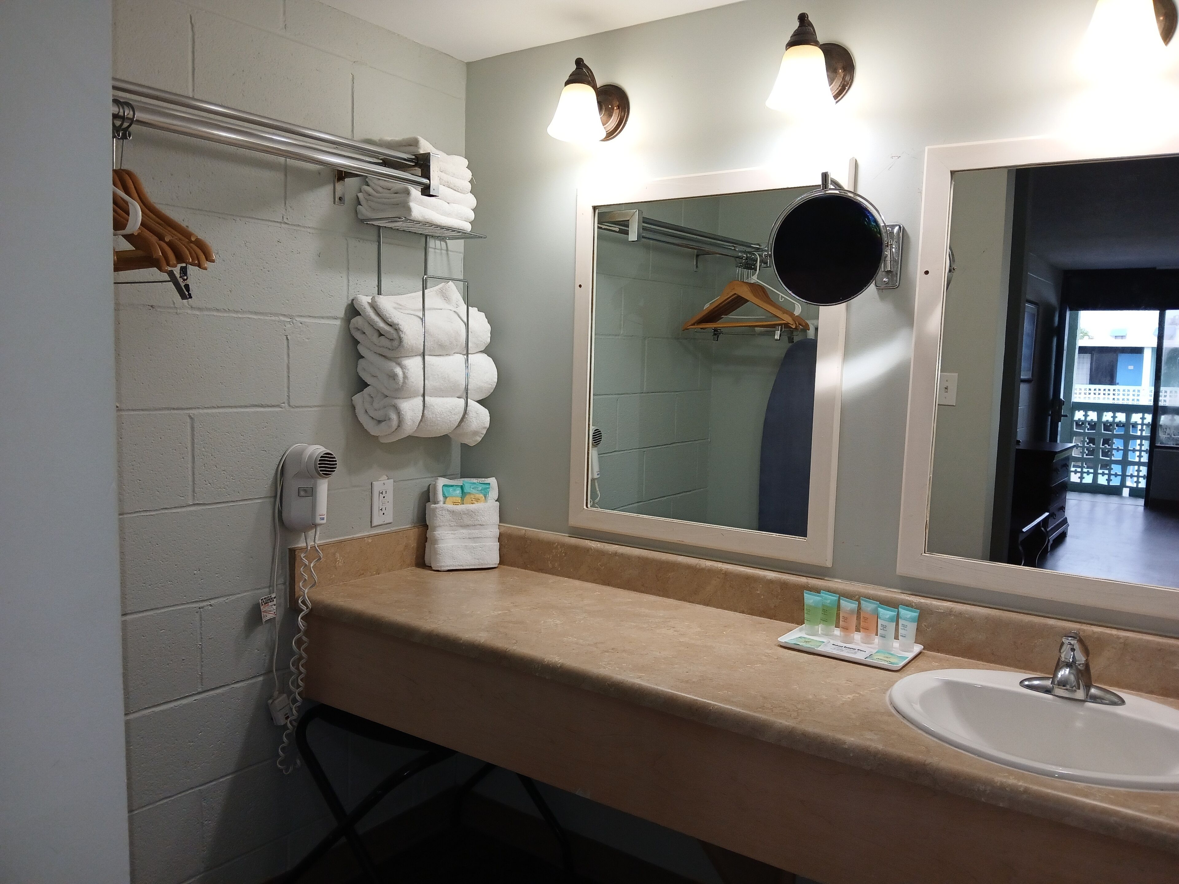 A bathroom with a beige countertop, a white sink, and a mirror with a round mirror and two wall-mounted lights. There are towels hanging on a rack and a hairdryer on the wall.