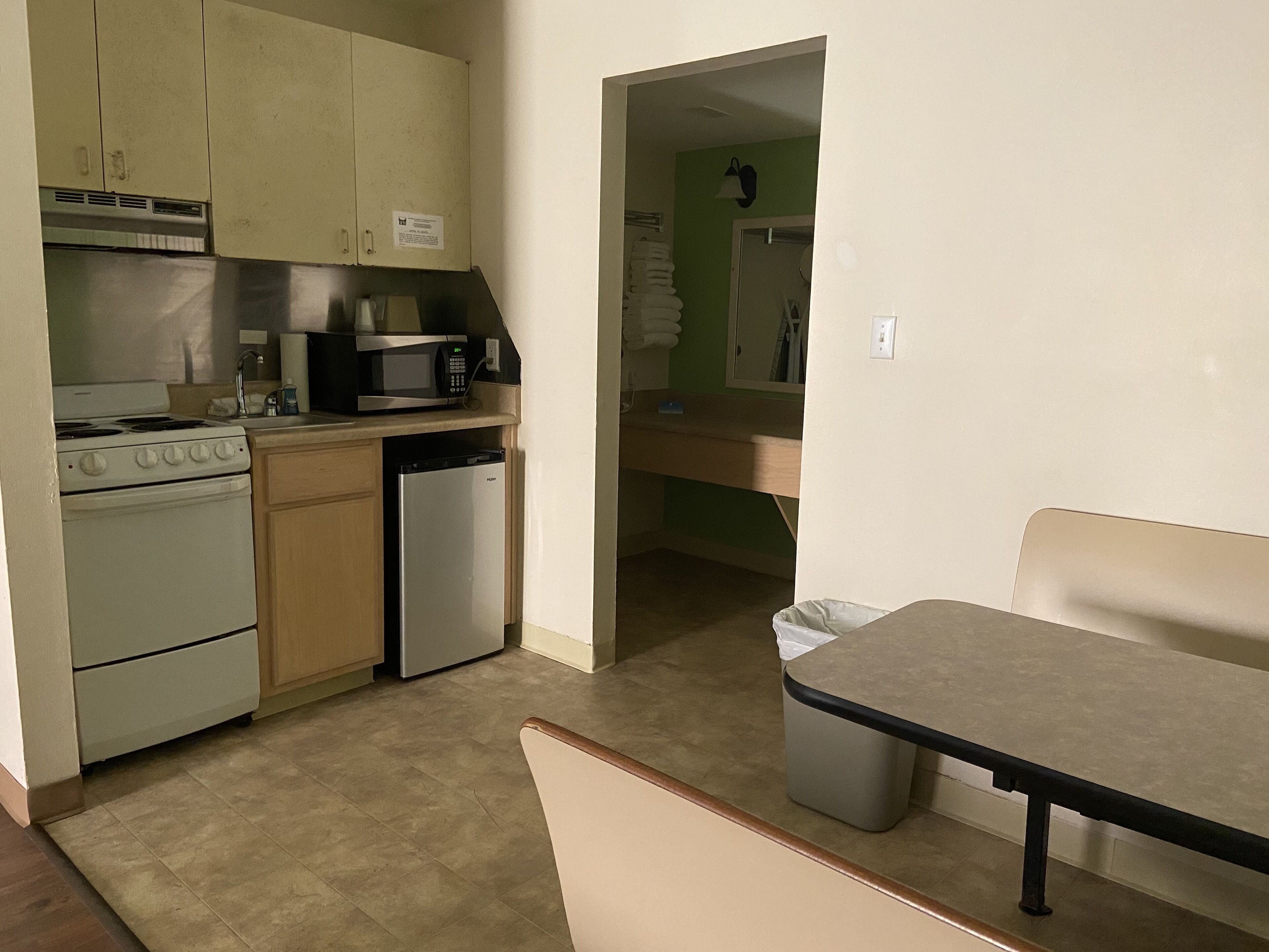 A compact kitchen with a stove, microwave, and dishwasher. There is a trash bin and a bench in the foreground.