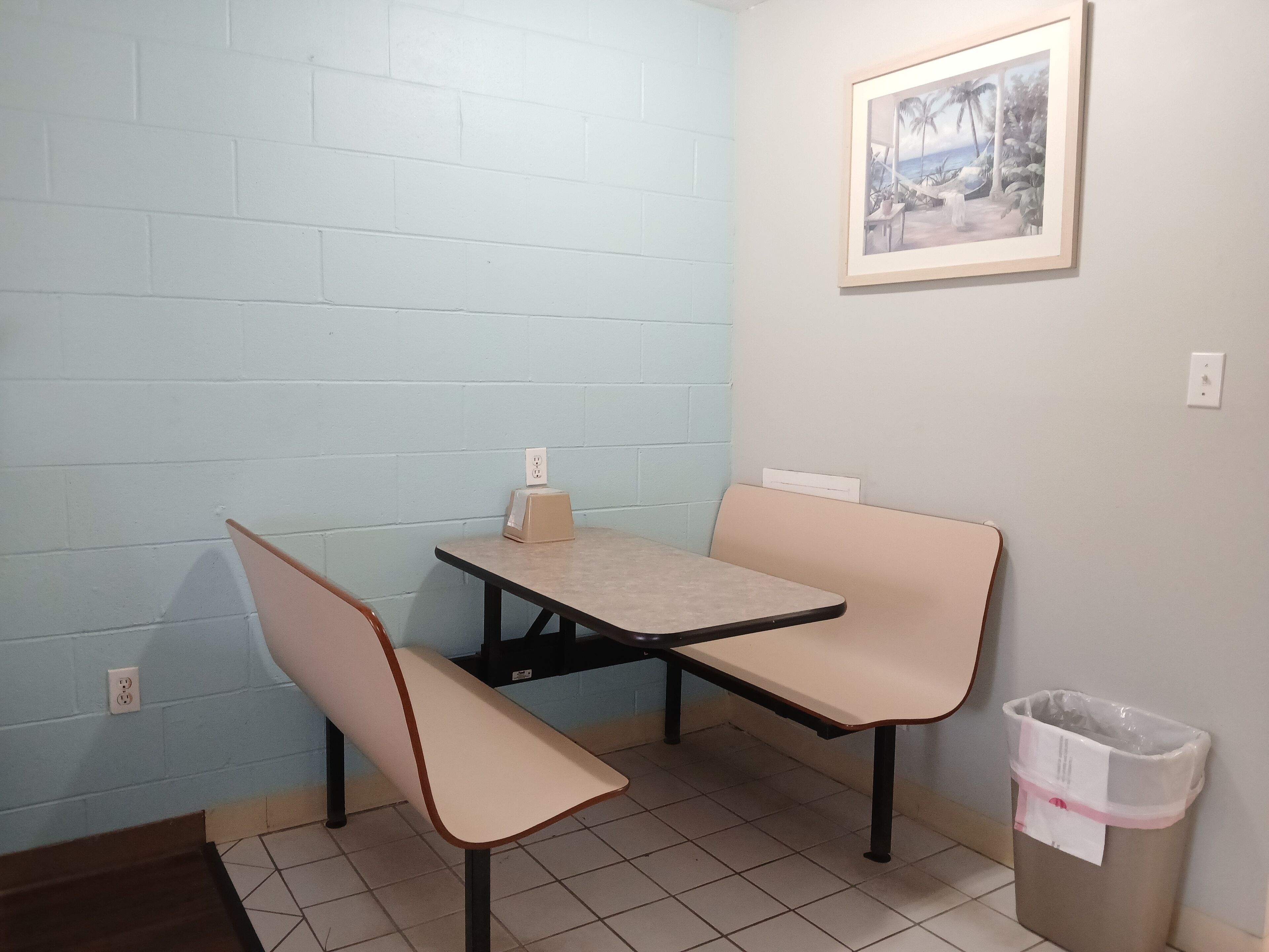 A small kitchenette with a table and bench, a trash bin, and a framed picture on the wall.