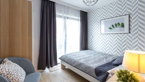Een moderne slaapkamer met een bed, een stoel, een nachtkastje met een lamp en een raam met gordijnen.