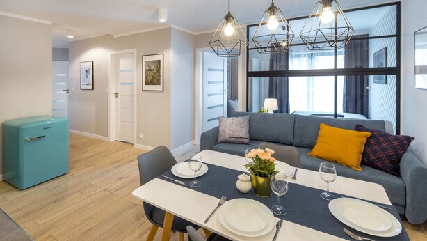 Een moderne woonkamer met een eettafel gedekt voor twee personen, een bank met kussens, een kleine koelkast en een groot raam met uitzicht.
