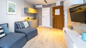 Een moderne woonkamer met een grijze bank, een flatscreen-televisie en een keuken met een houten kast en een witte deur.