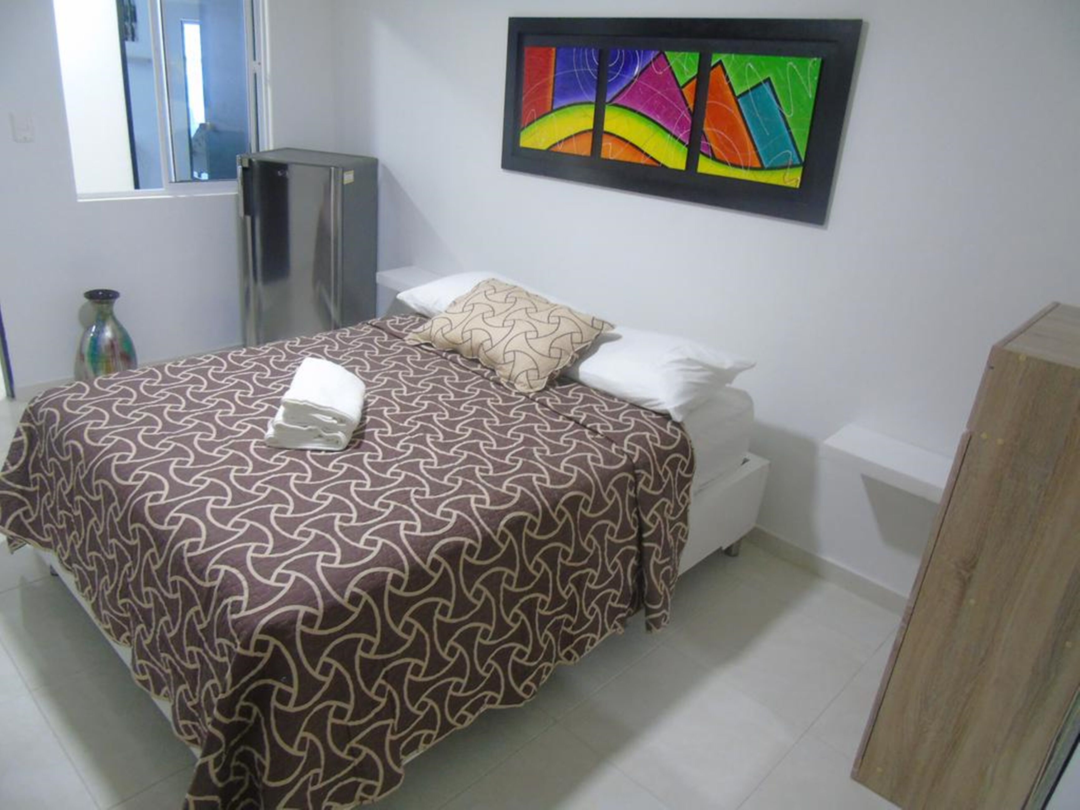 A hotel room with a bed, a small cabinet, a window, and a colorful abstract painting on the wall.