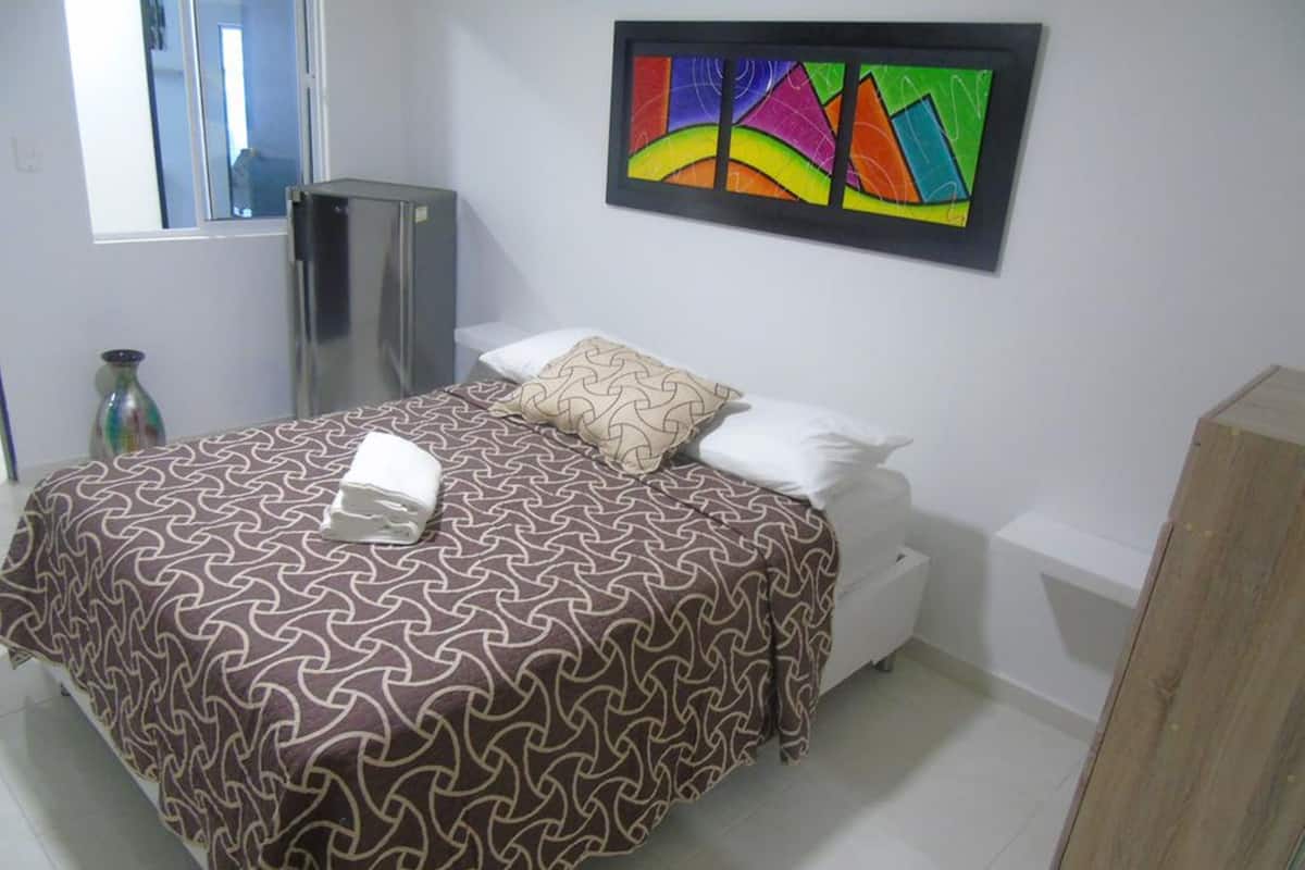 A hotel room with a bed, a small cabinet, a window, and a colorful abstract painting on the wall.