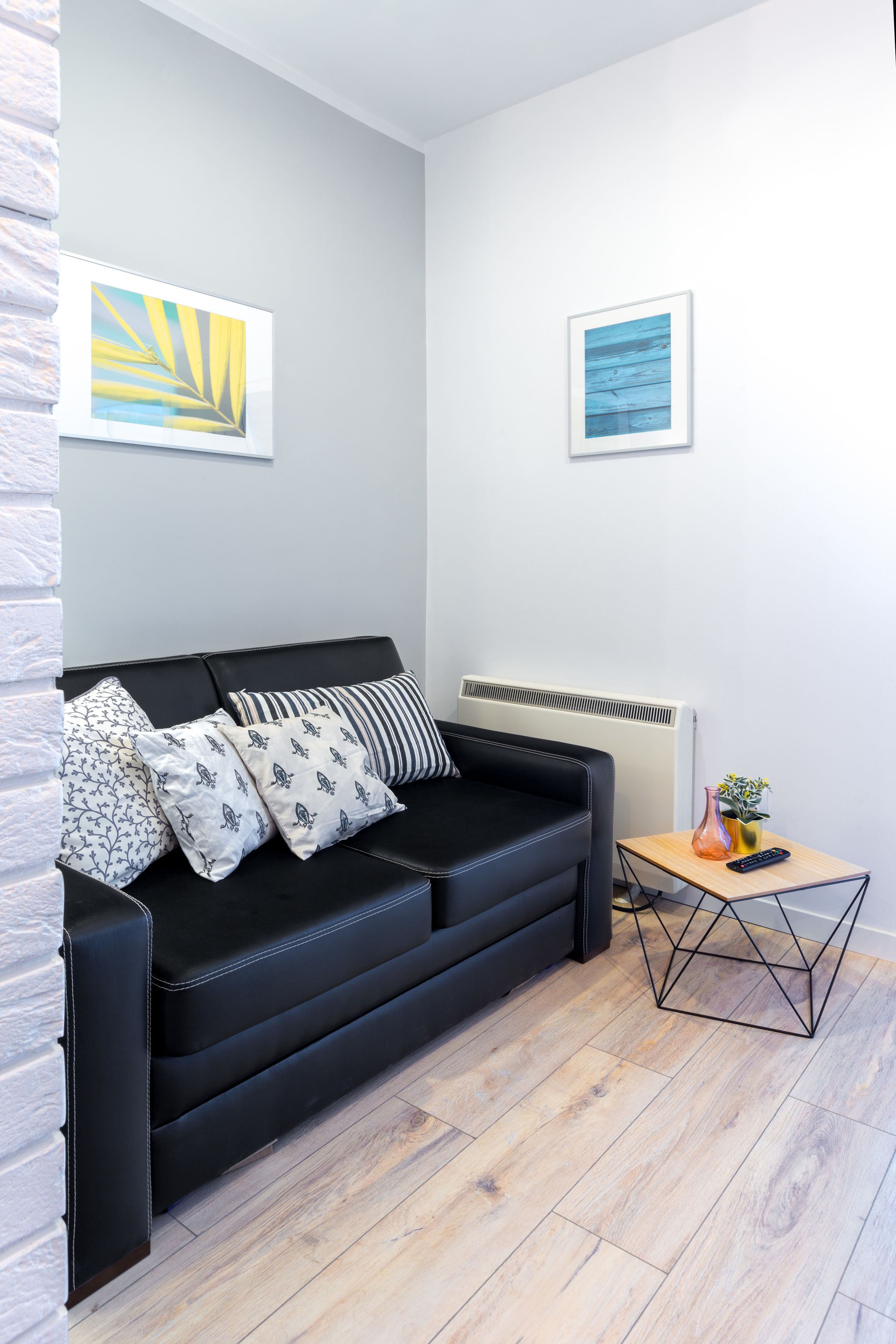 A modern living room with a black leather sofa, patterned cushions, a small wooden side table with a vase and remote, and two framed artworks on the wall.