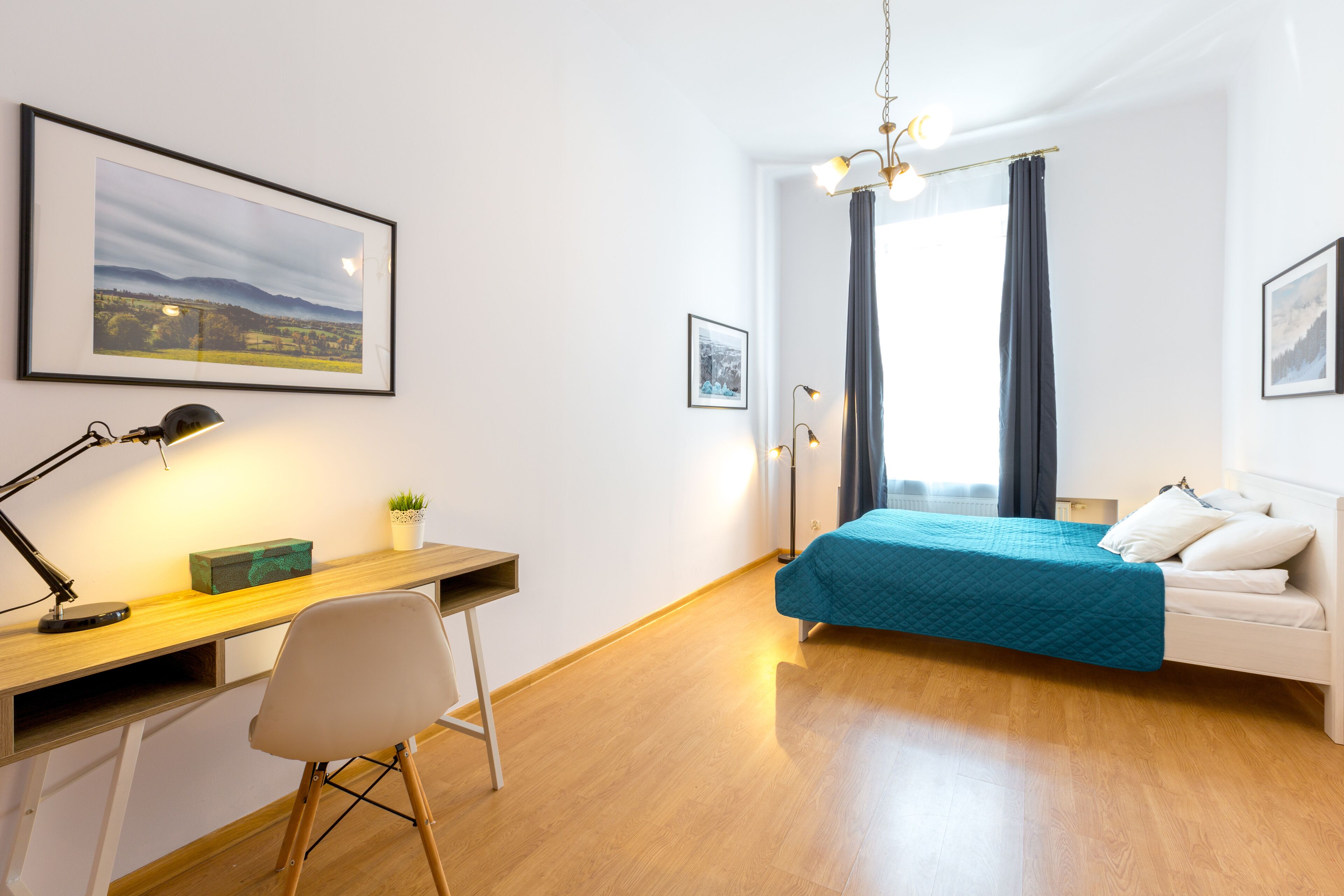 A bedroom with a wooden desk, a chair, a bed with a teal bedspread, a white sofa, and a large window with curtains.