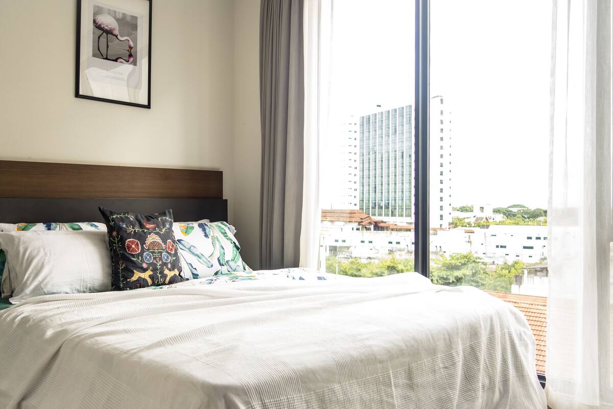 A hotel room with a large bed, a nightstand, a lamp, and a view of the city through large windows.