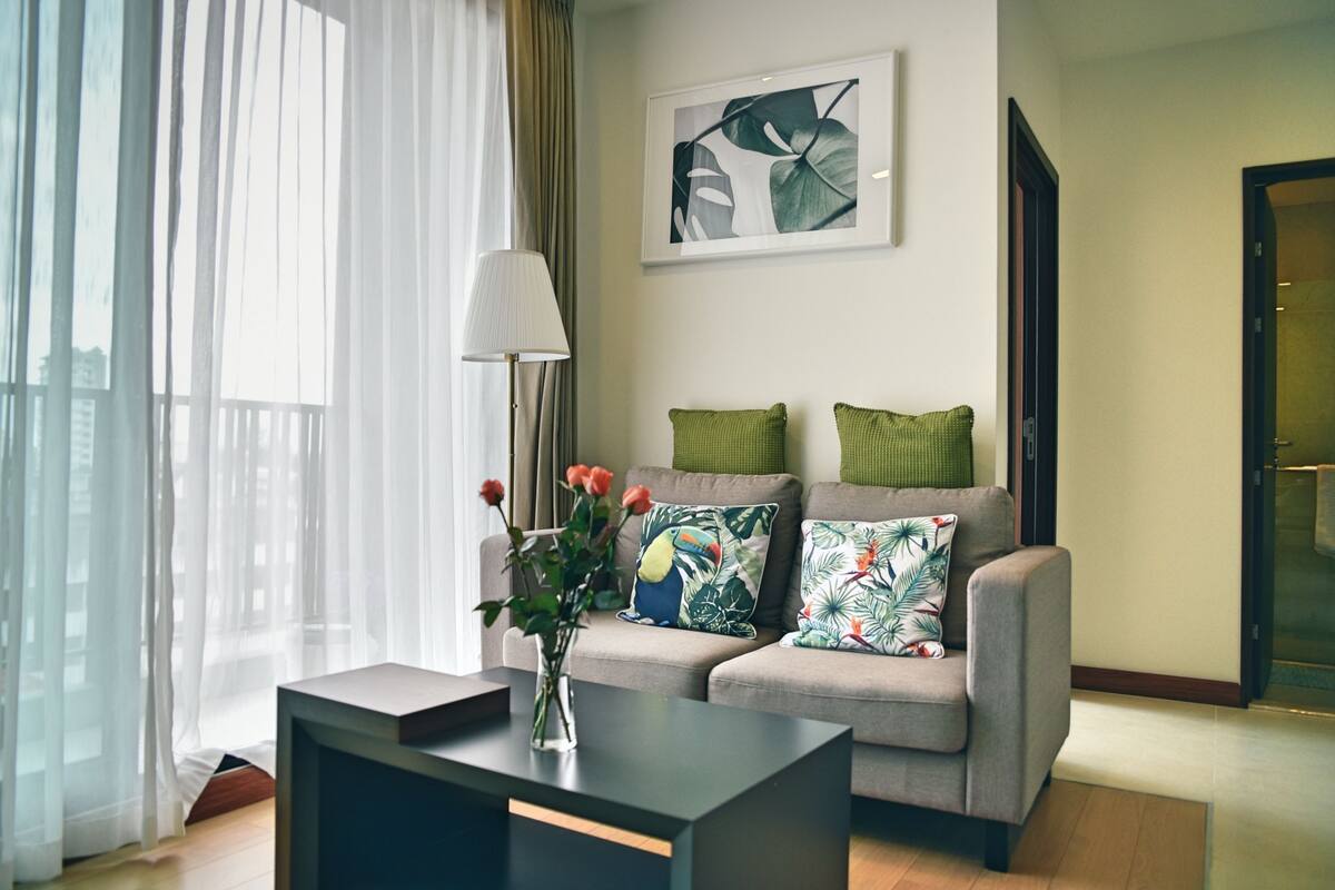 A modern living room with a grey sofa, green pillows, a vase with flowers, and a framed artwork on the wall.