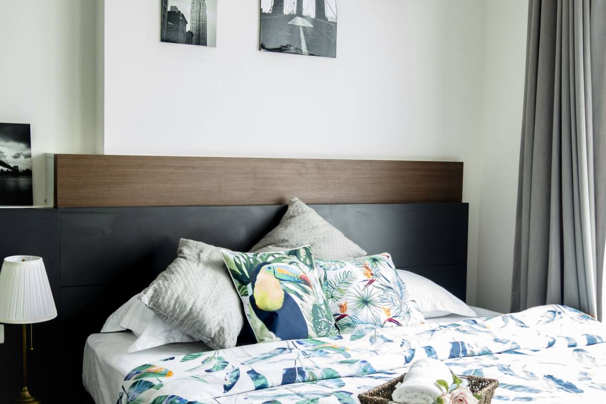 A neatly made bed with a tropical leaf pattern bedspread, a wooden headboard, and a nightstand with a lamp. There are three framed pictures on the wall above the bed.