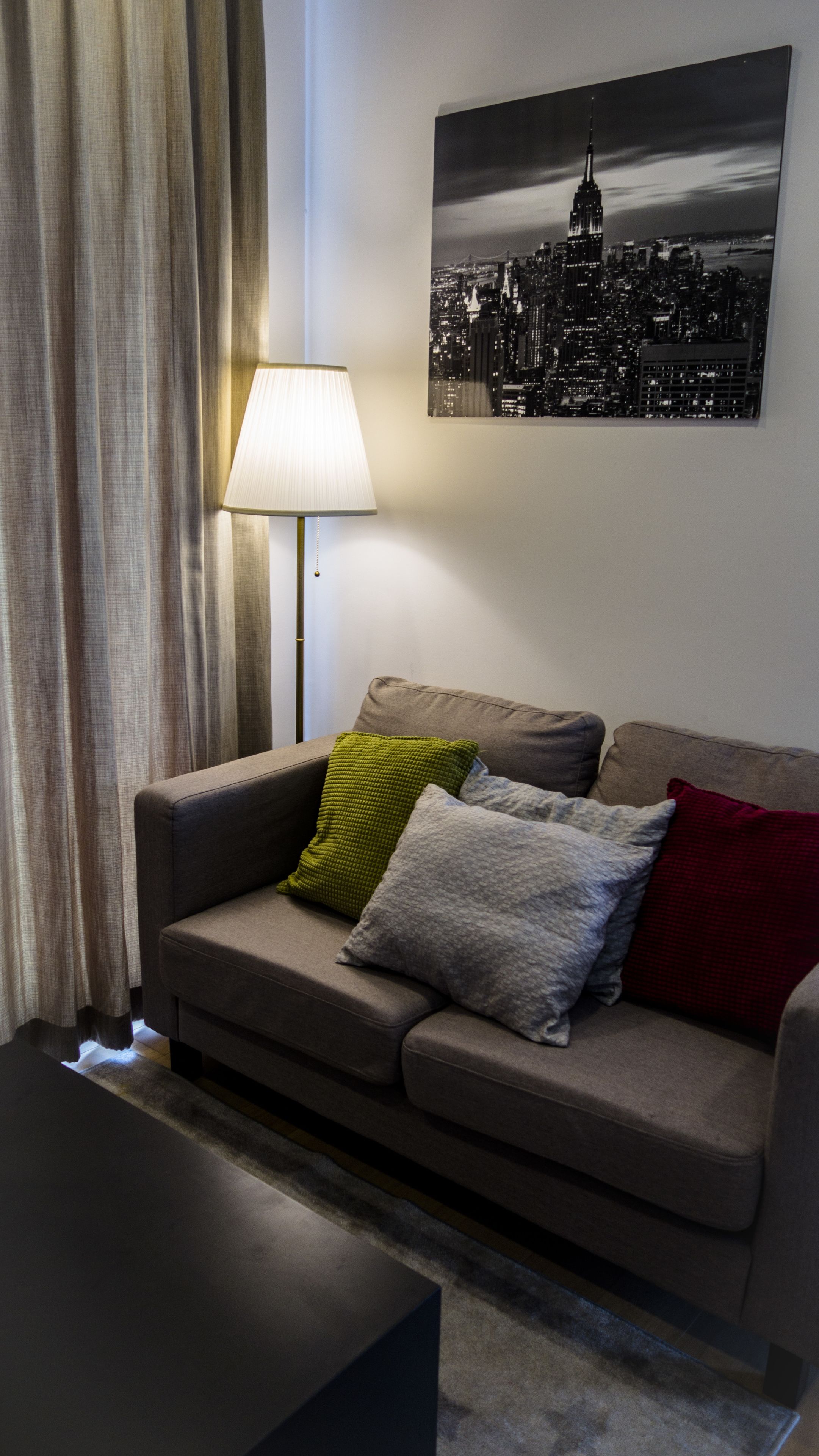 Ett vardagsrum med en soffa, en lampa och en tavla som föreställer en stadsbild på väggen.