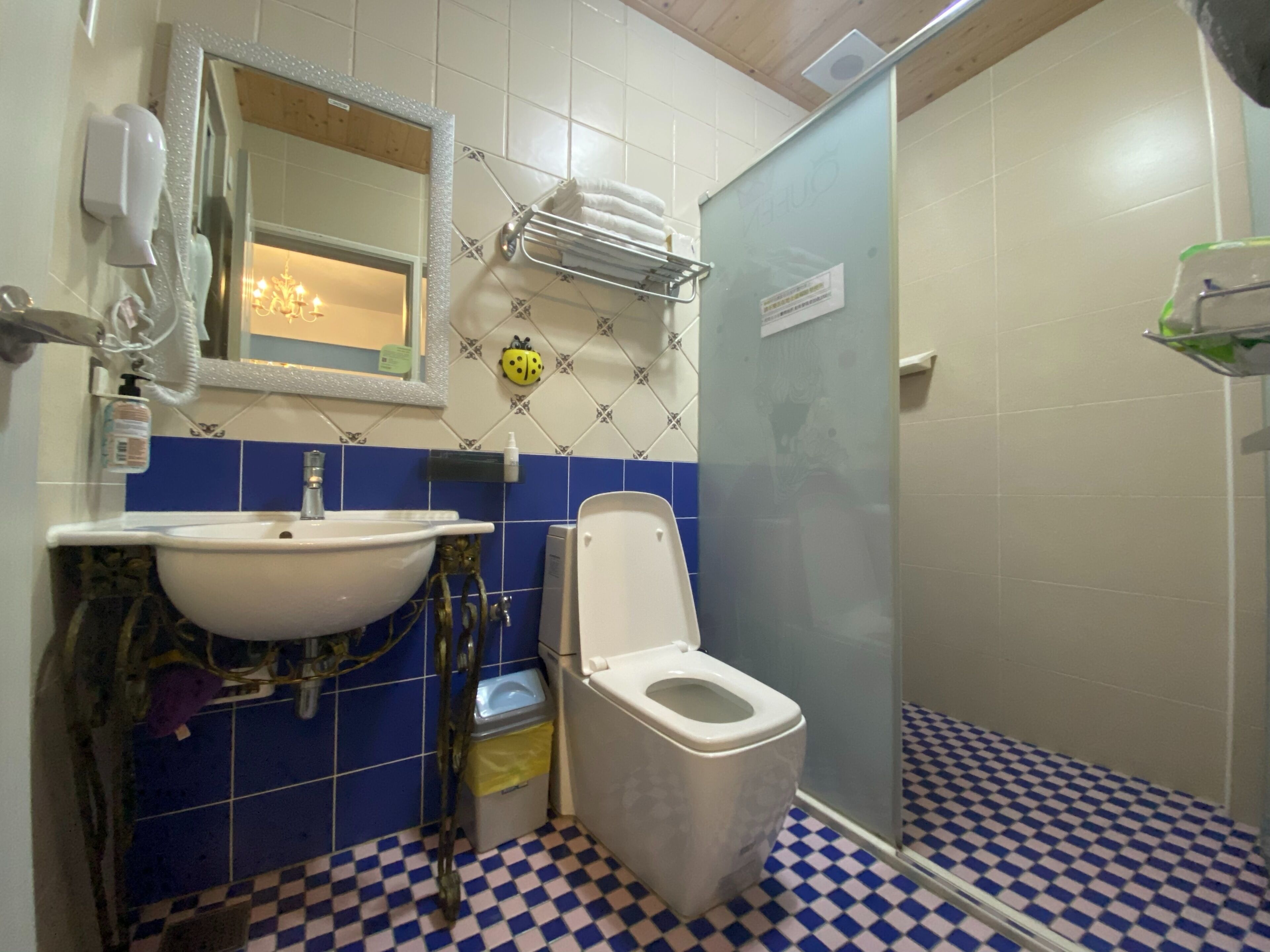 A bathroom with a white sink, toilet, and shower. There is a mirror above the sink, and a towel rack is mounted on the wall. The floor has a blue and white checkered pattern.
