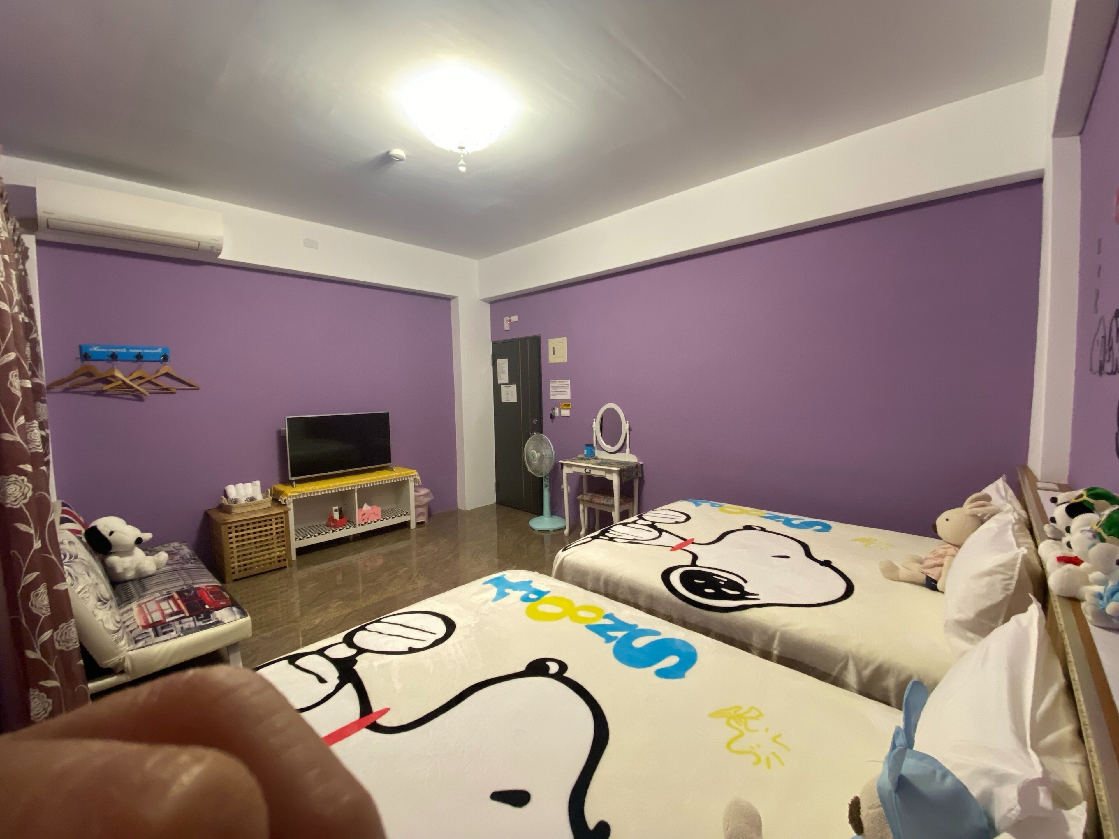 A room with purple walls, a bed with a cartoon character design, a TV, a fan, and a small table with a mirror.