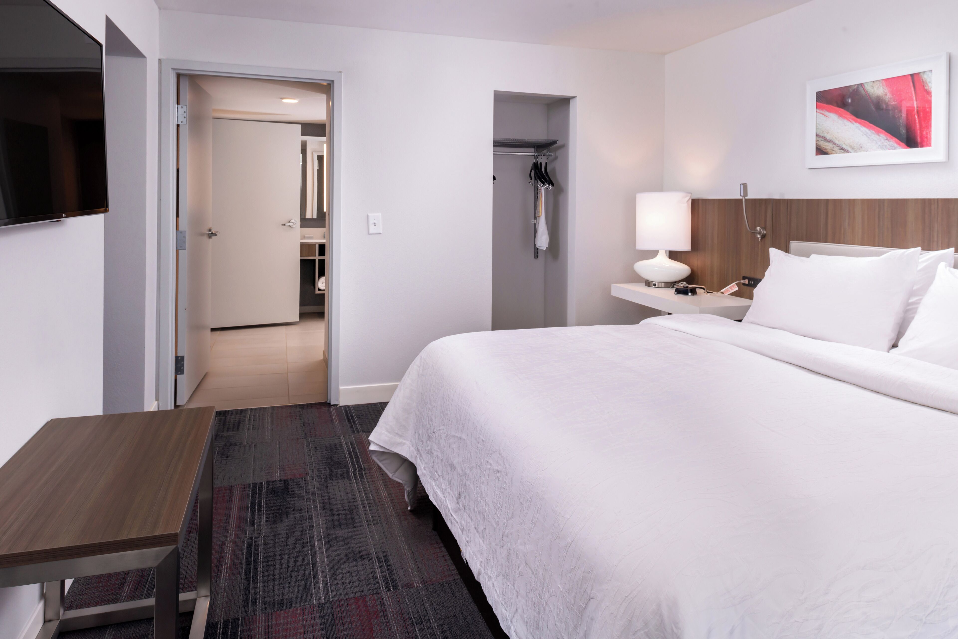 Een hotelkamer met een bed, nachtkastje, lamp en uitzicht op een gang met een kapstok.