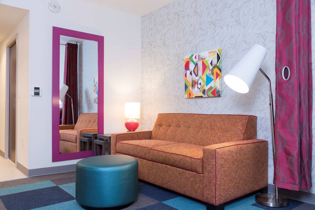 A modern hotel room with a sofa, ottoman, lamp, and a colorful abstract painting on the wall.