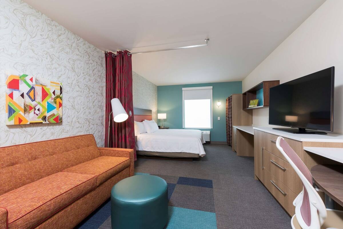 A hotel room with a bed, a sofa, a television, a desk, and a colorful abstract painting.