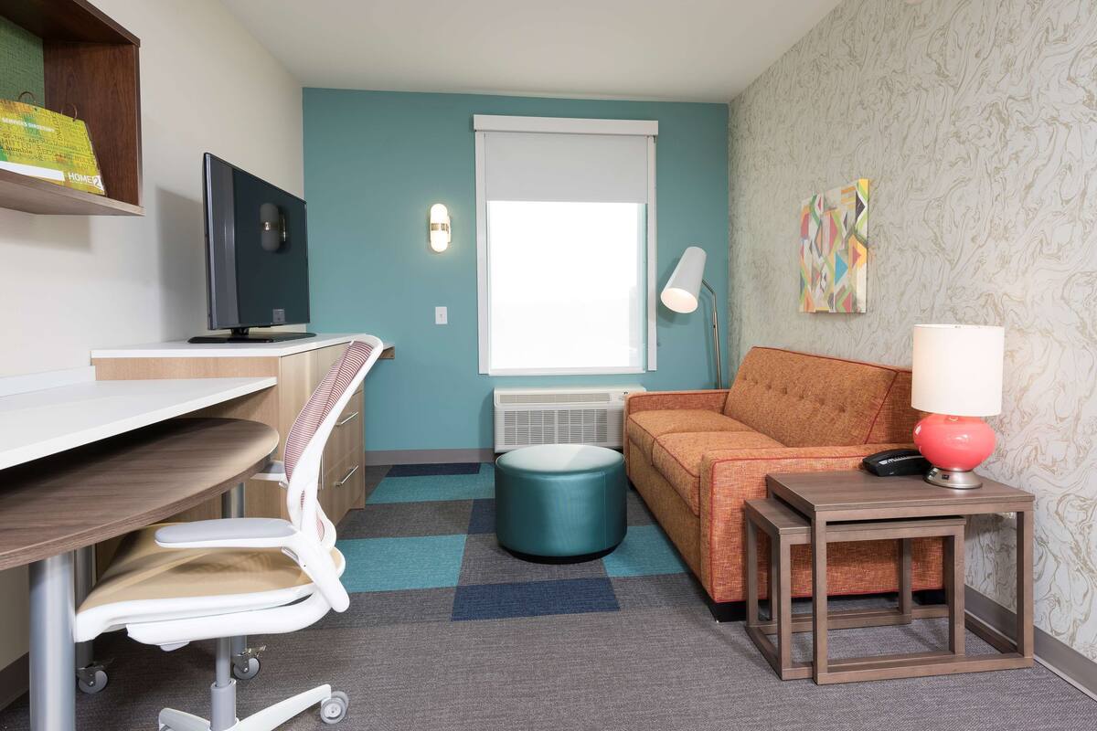 A compact room with a desk, a chair, a sofa, a small table with a lamp, and a window with blinds.
