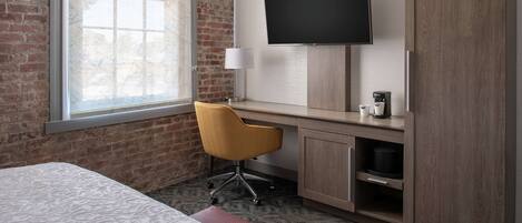 A hotel room with a bed, a desk with a chair, a TV, a lamp, and a coffee machine.