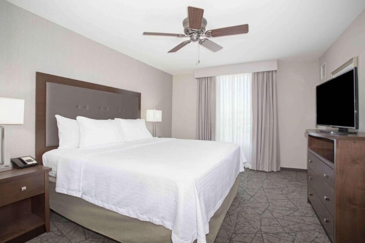 A hotel room with a bed, a TV, a desk, a phone, a ceiling fan, and a window with curtains.