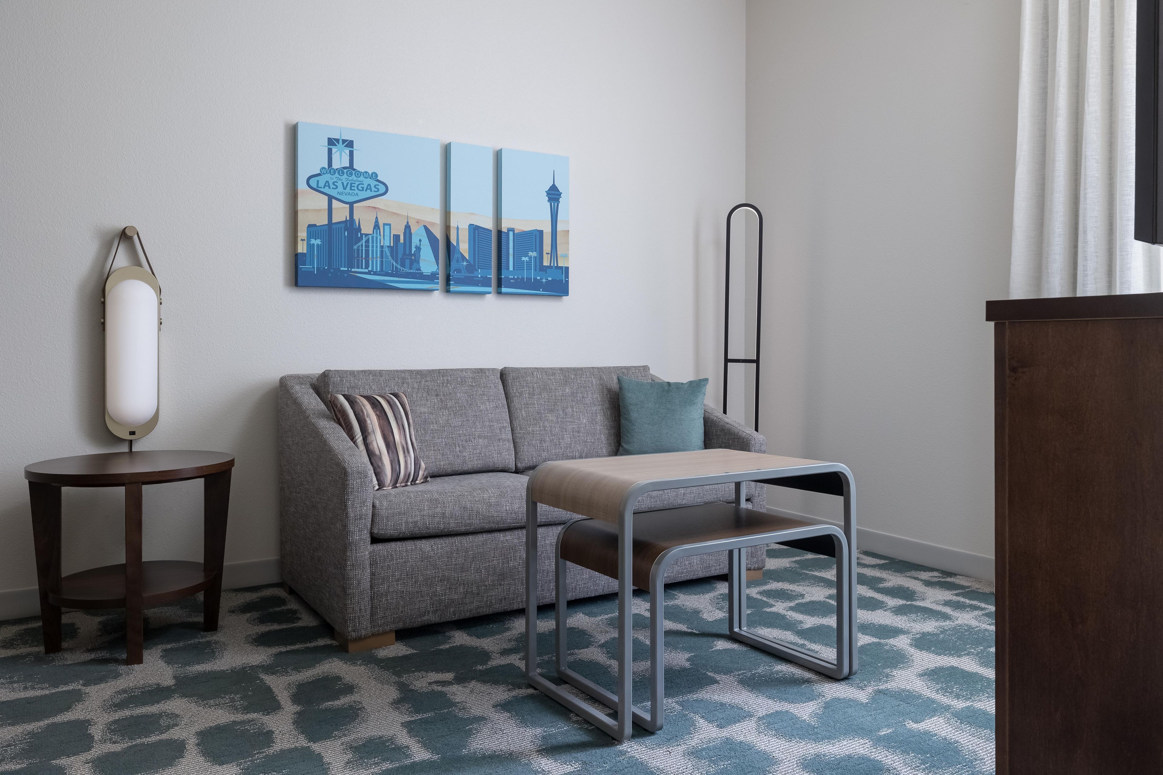 Une chambre d’hôtel avec un canapé gris, une petite table, une lampe et un tableau représentant un paysage urbain accroché au mur.