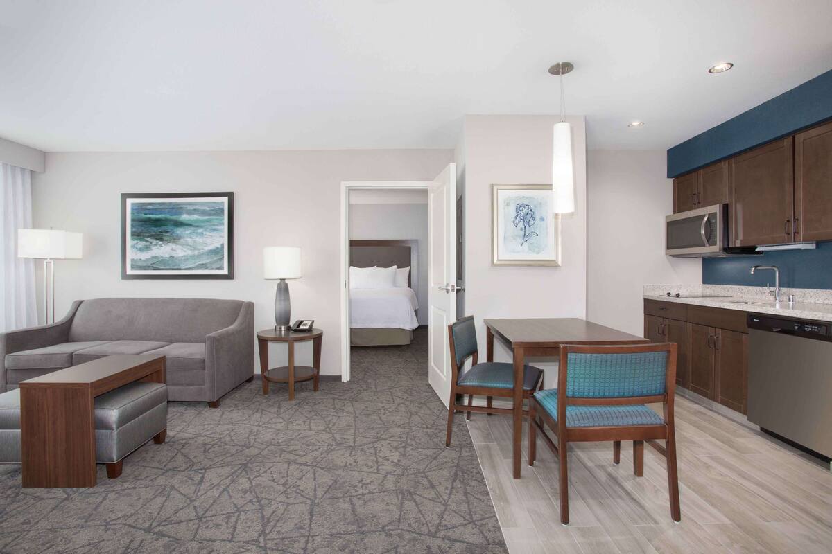 Una habitación de hotel con un sofá gris, una mesa de comedor con dos sillas, una cama con ropa de cama blanca, una zona de cocina con microondas y un cuadro en la pared.