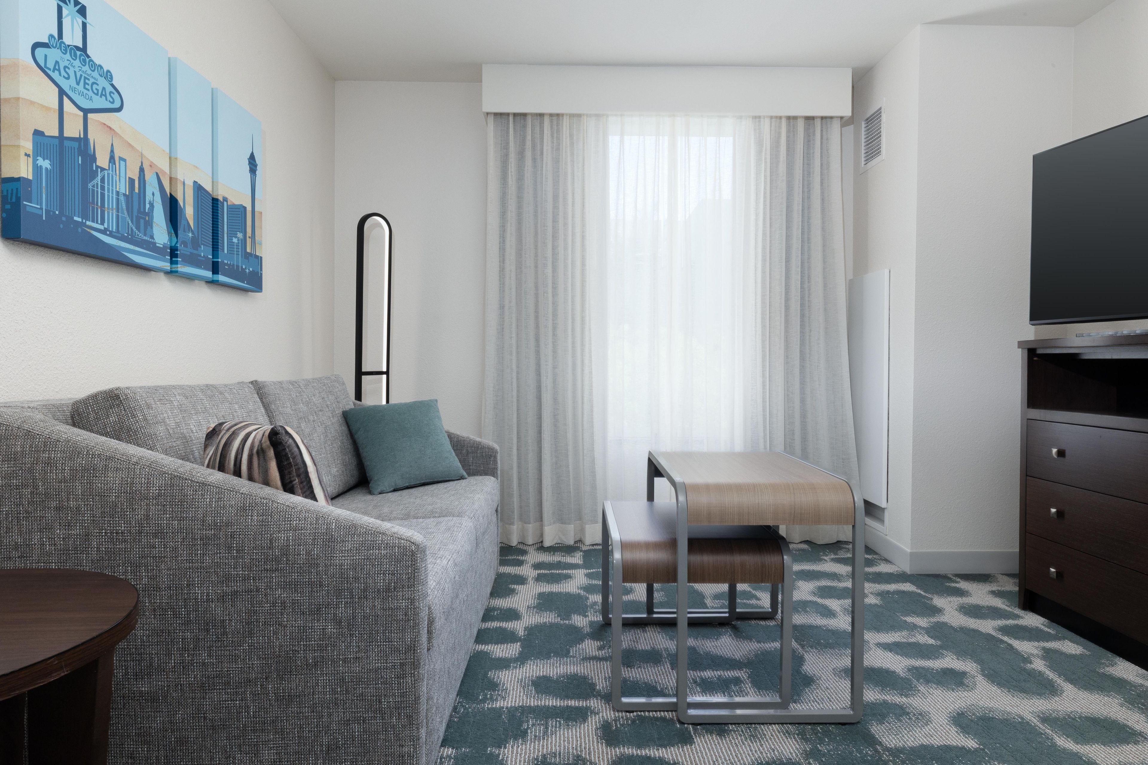 Habitación de hotel con sofá, una mesita, televisor y un cuadro de un paisaje urbano.