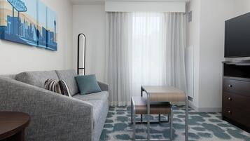 Habitación de hotel con sofá, una mesita, televisor y un cuadro de un paisaje urbano.