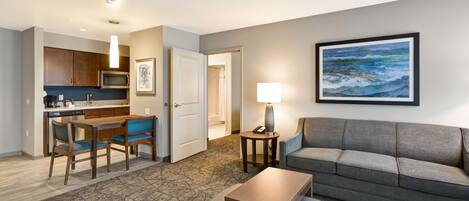 A modern hotel room with a kitchenette, a dining area, a sofa, and a painting on the wall.