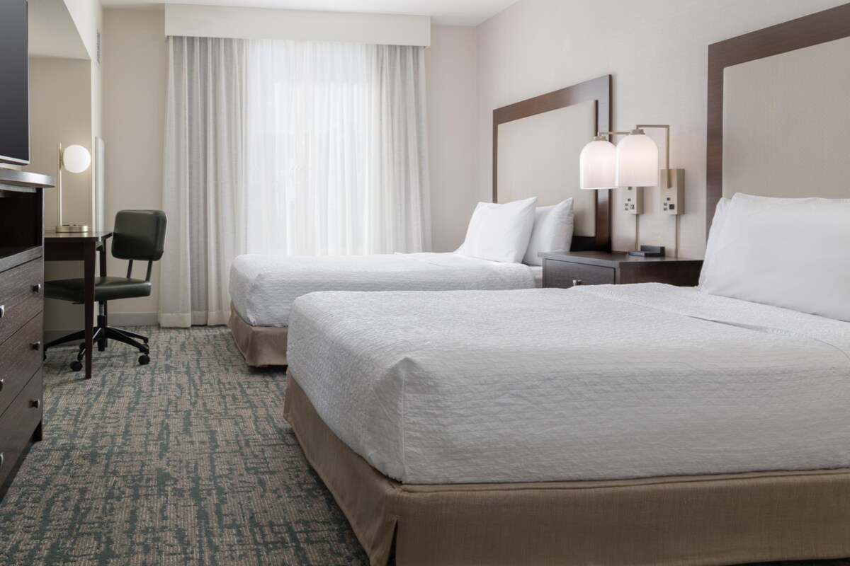 Une chambre d’hôtel avec deux lits, un bureau, une chaise, une commode et une fenêtre avec des rideaux.