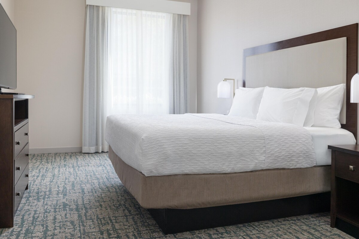 Une chambre d’hôtel avec un lit, une table de chevet, une lampe, une commode et une fenêtre avec des rideaux.