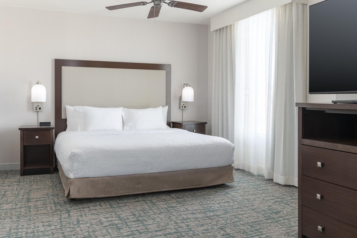 Une chambre d’hôtel avec un grand lit, deux tables de chevet, un ventilateur de plafond, un téléviseur à écran plat et une grande fenêtre avec des rideaux.