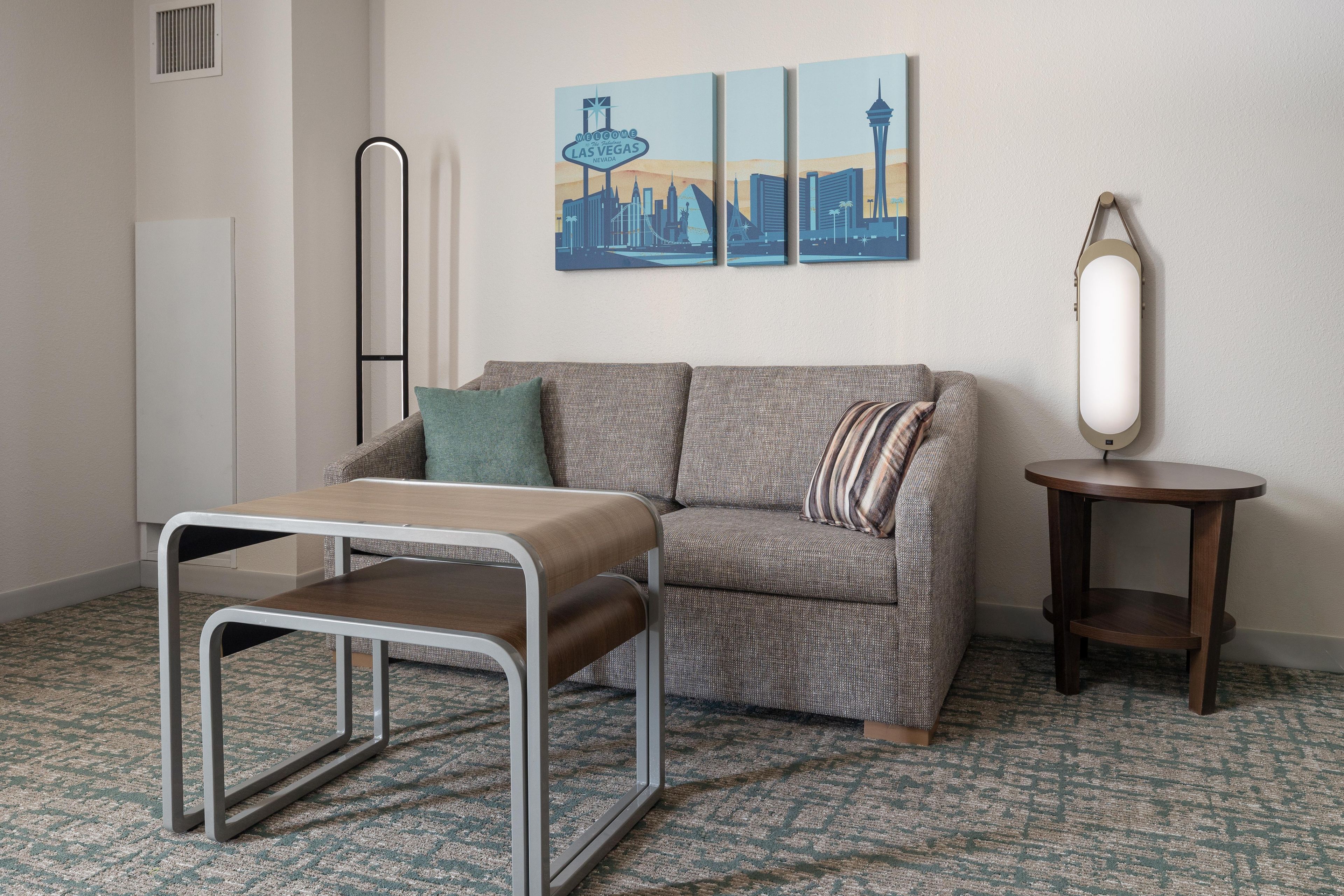 A room with a patterned carpet, a sofa with cushions, a small table, a wall-mounted mirror, and a framed cityscape painting.
