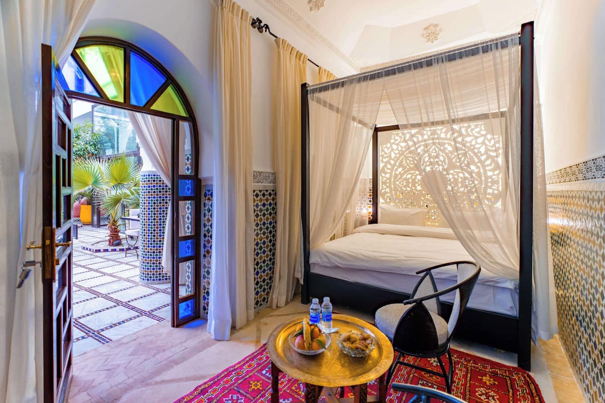 A hotel room with a bed, a small table with snacks, a chair, and a view of the courtyard.