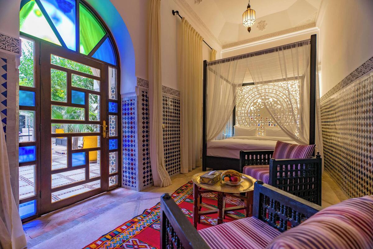 A room with a bed, a window with a view, a table with a basket, and patterned walls.