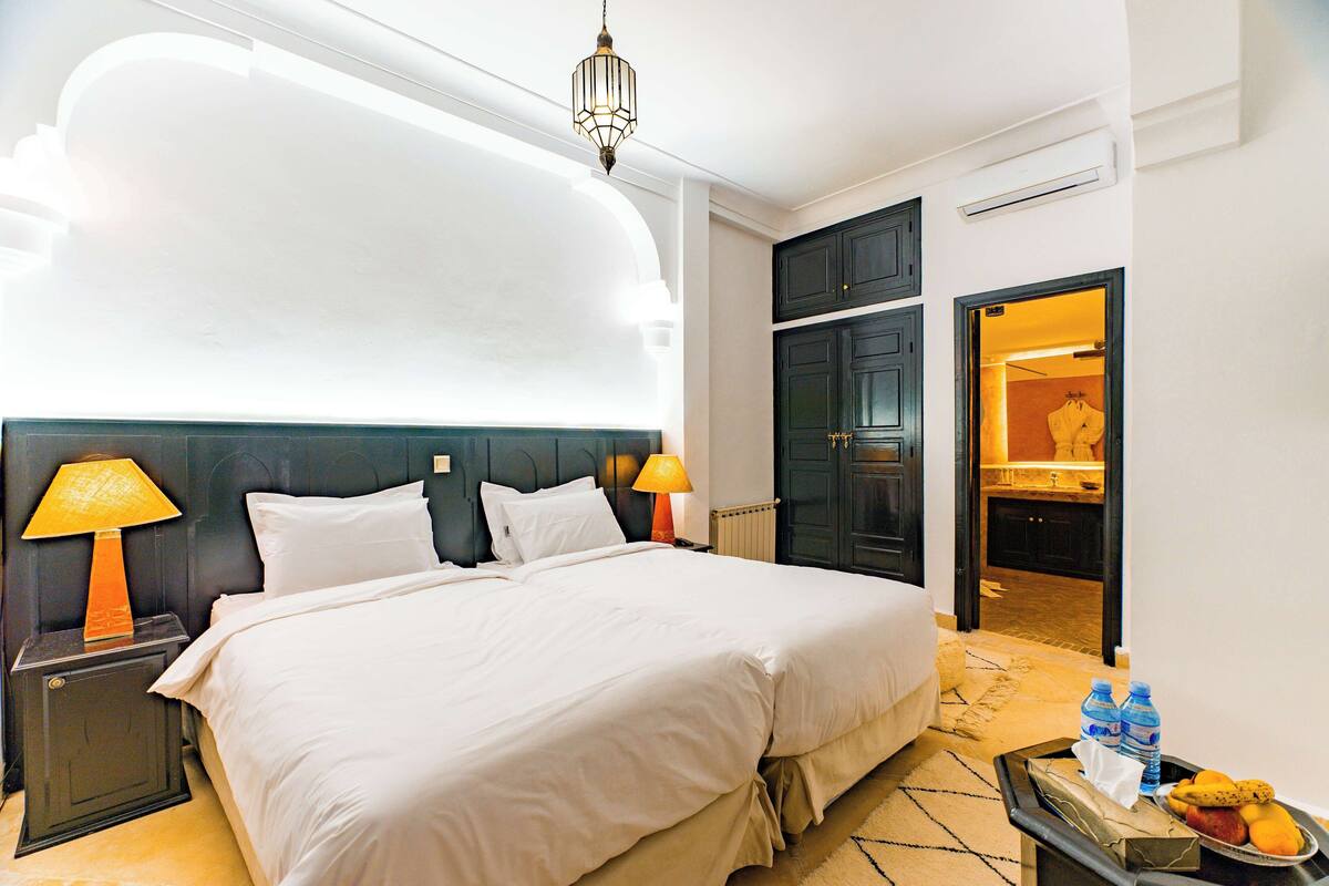 A hotel room with a large bed, two bedside lamps, a nightstand, a tray with fruits and water bottles, and a view of the bathroom through an open door.