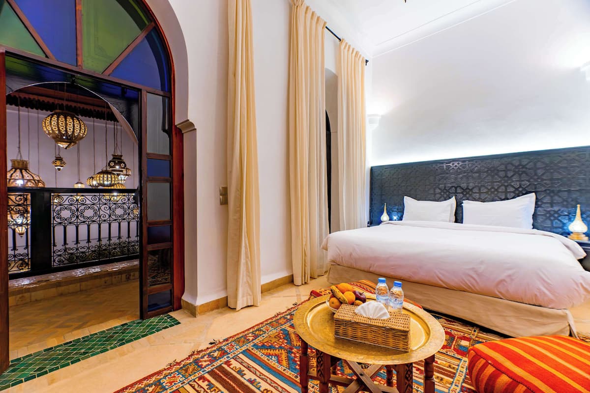A hotel room with a large bed, a small round table with fruits and a bottle, a colorful rug, and a decorative wall with hanging lights.