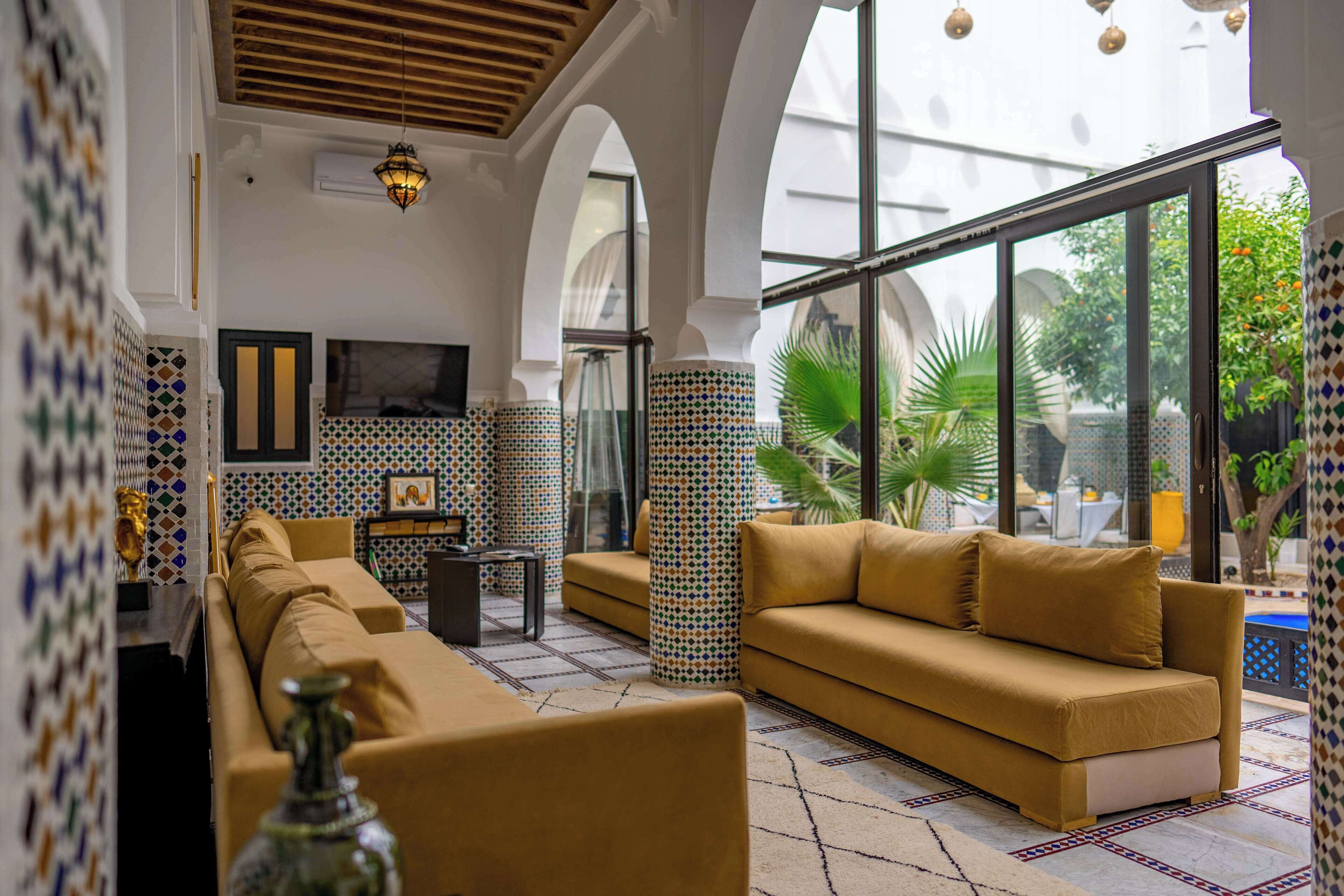 A modern living room with a sofa, a coffee table, and a TV. The room has patterned tiles on the walls and a large window with a view of greenery.