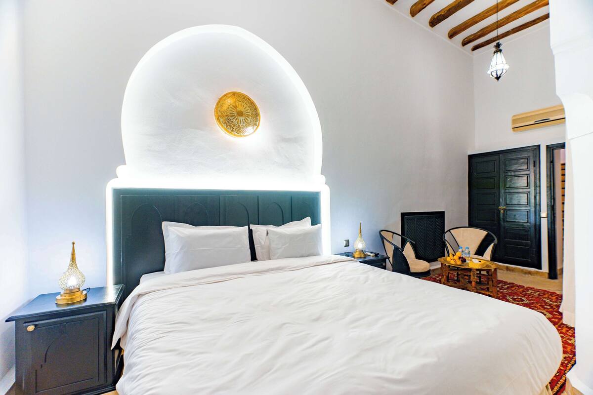 A hotel room with a large bed, a nightstand with a lamp, a chair, and a small table with a bottle.