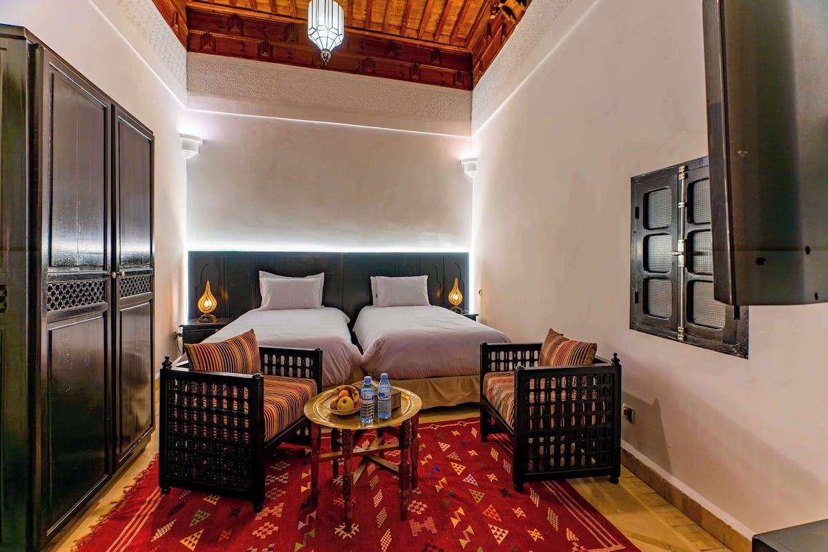 A hotel room with two beds, a small table with water bottles, and a patterned red carpet.