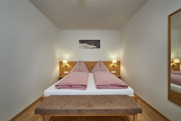 A double bed with checkered bedding, two bedside lamps, a small painting on the wall, and a wooden bench at the foot of the bed.
