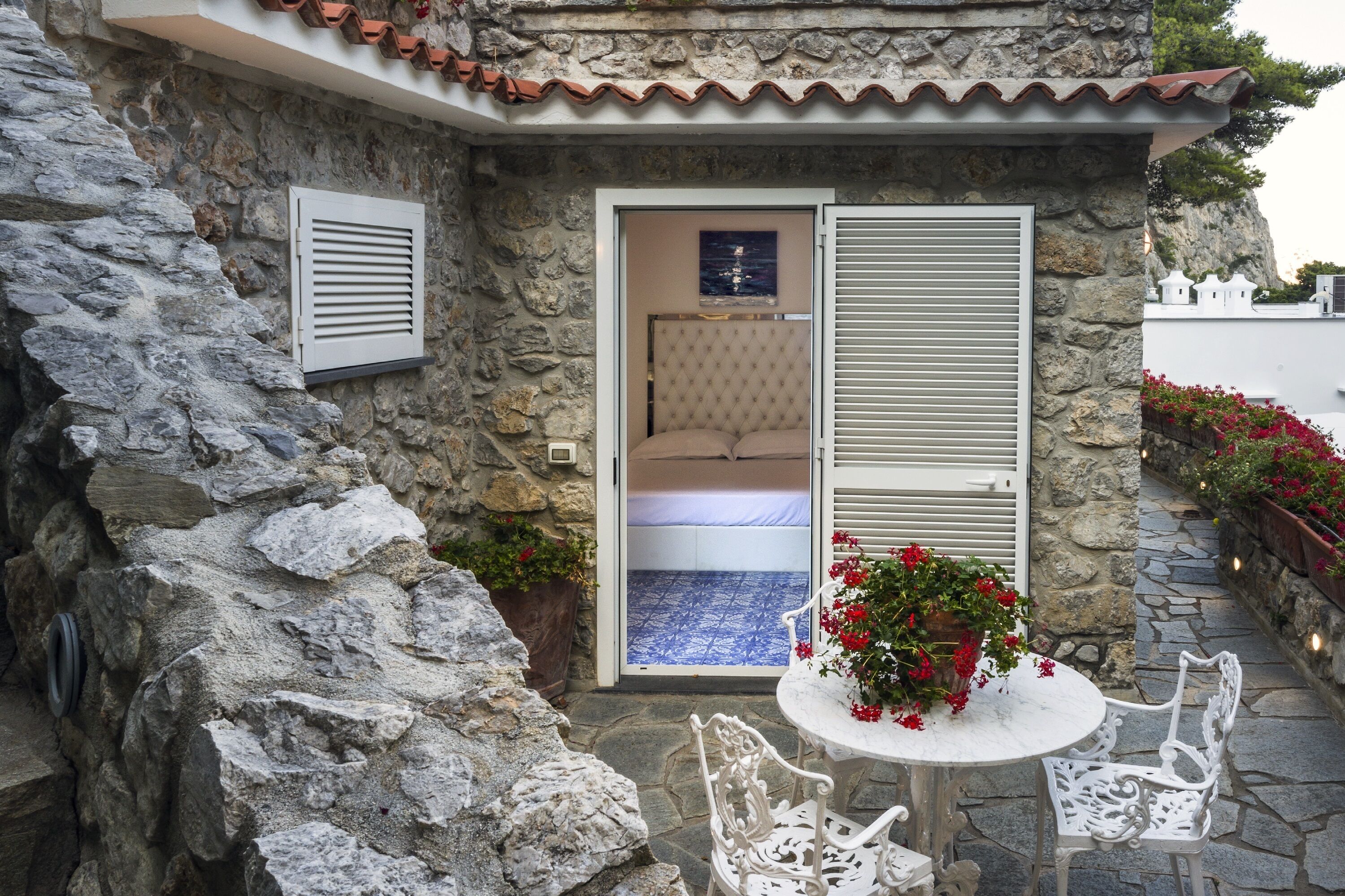 Edificio de piedra con un dormitorio que tiene una cama y vistas a través de una puerta. Hay una mesa con sillas y plantas en macetas afuera.