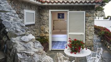 Edificio de piedra con un dormitorio que tiene una cama y vistas a través de una puerta. Hay una mesa con sillas y plantas en macetas afuera.