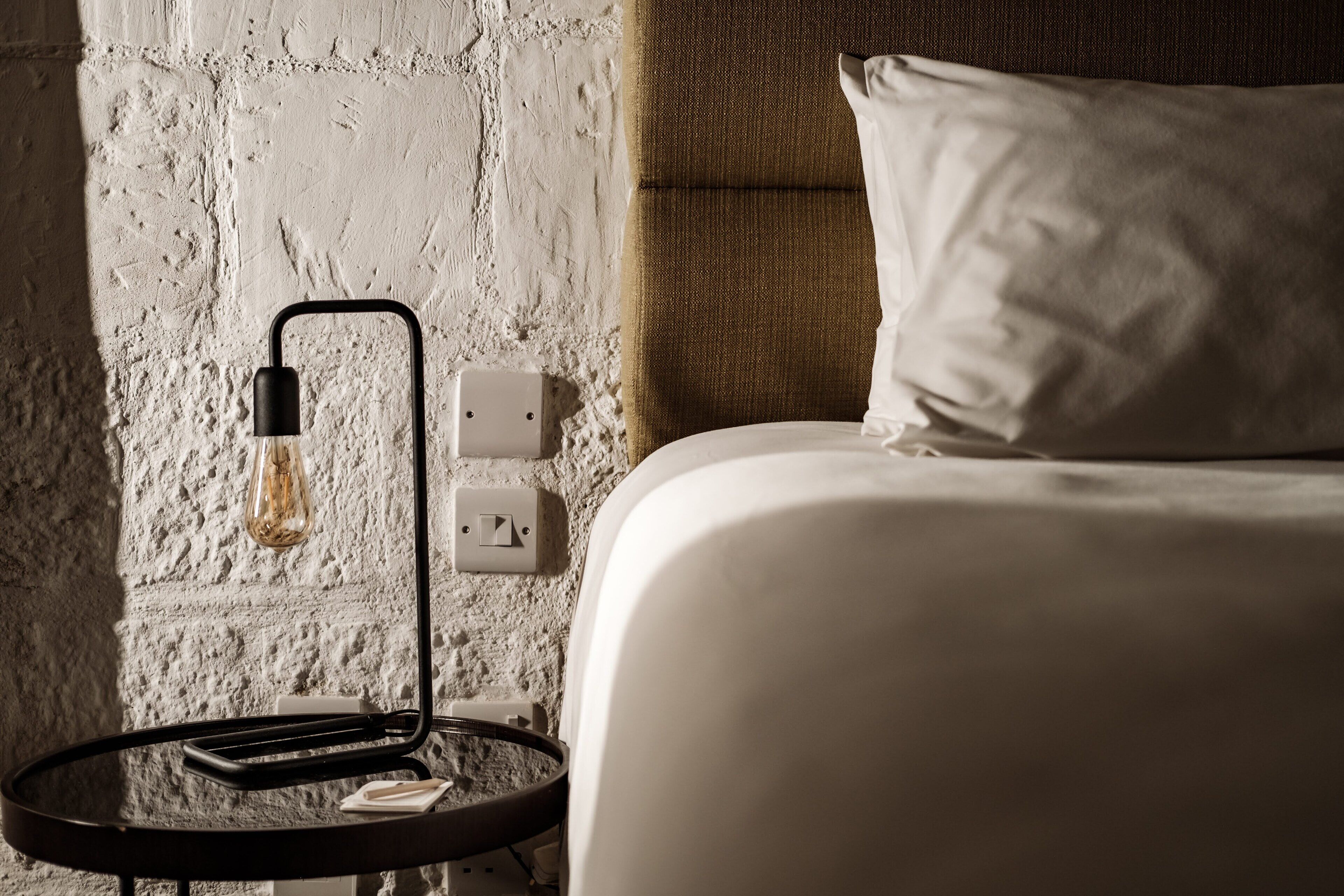 A bed with white bedding, a round nightstand with a black lamp, and a textured white wall.
