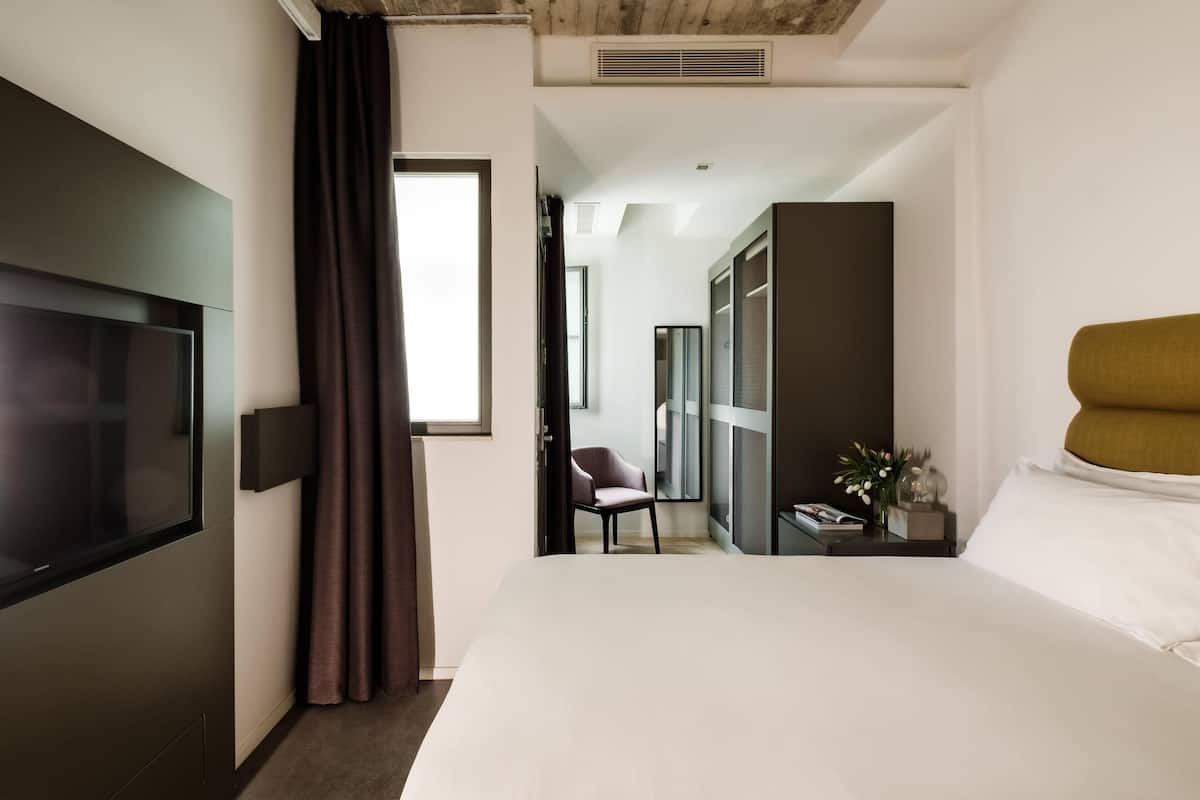 A modern hotel room with a large bed, a flat-screen TV, a chair, a small table with a vase, and a wardrobe.