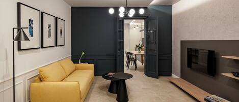 Una sala de estar moderna con un sofá amarillo, una mesa de centro negra y un televisor en la pared. Hay cuadros enmarcados en la pared y una lámpara en el techo.